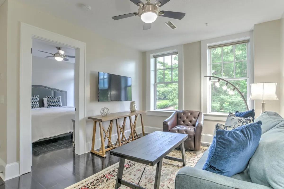 The image showcases a sunlit living area with a plush, comfortable sofa and a stylish armchair arranged around a sleek wooden coffee table. Large windows flood the space with natural light, while warm lighting creates a welcoming ambiance for cozy evenings. A modern TV is mounted on the wall, and the overall decor is inviting and thoughtfully designed.