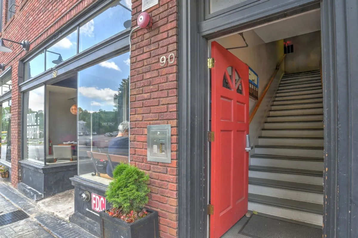 This image features the exterior entrance to the building, showcasing a classic red door, an ascending staircase, large windows, and a brick façade accented with a vibrant planter box. The visual gives a direct sense of arrival, emphasizing the building's historic charm and the welcoming atmosphere of the entryway.
