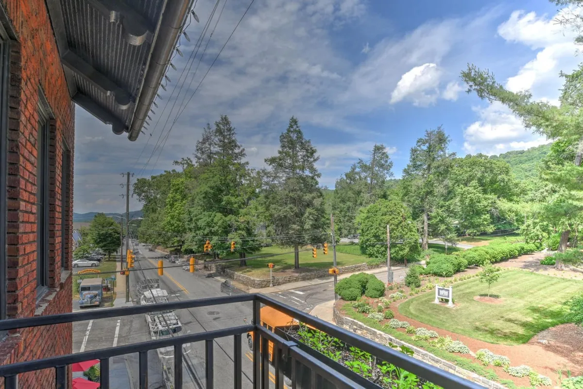 A serene view from a balcony looking out over lush greenery, distant mountains, and a city street below. The scene invites relaxation, morning coffee rituals, and people-watching while staying connected to the lively neighborhood.