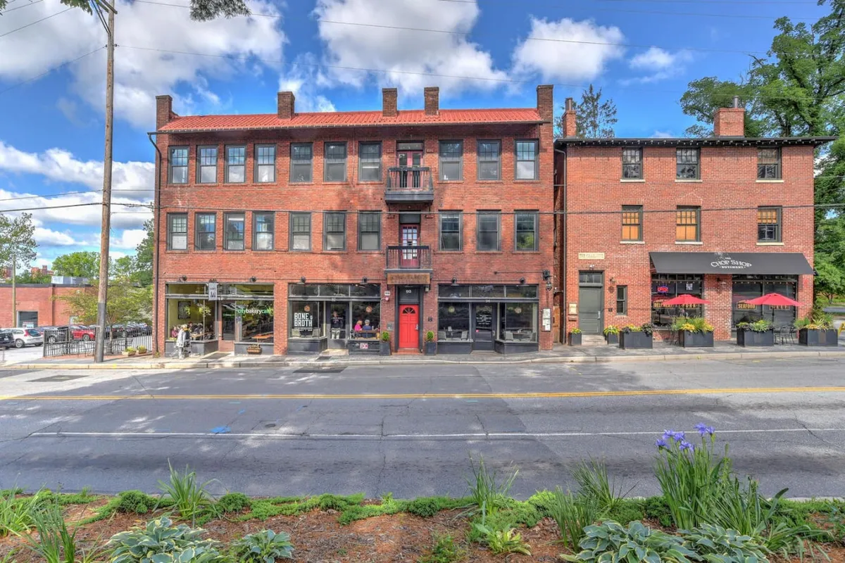 A lively streetscape featuring a red door, balcony, and a row of charming storefronts with visible signage. The scene captures people strolling, shopping, and dining outdoors, evoking the vibrant community atmosphere of the neighborhood.