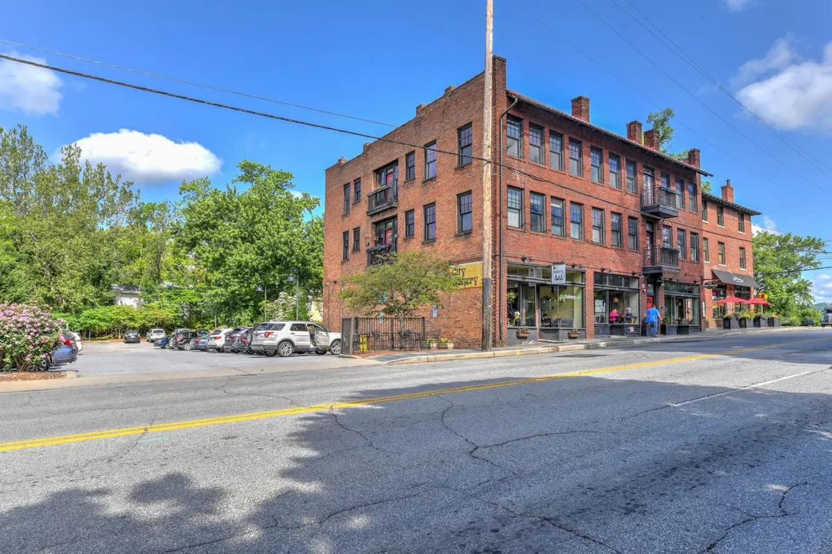 A historic brick building framed by green trees and sidewalk storefronts. The scene is alive with people shopping, dining, and engaging in conversation, highlighting the area’s strong sense of community and charm.