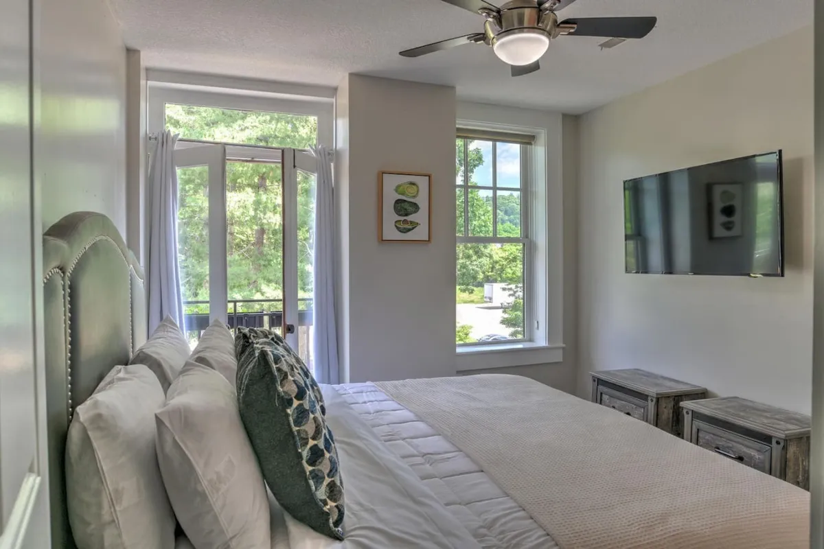 A relaxing bedroom with a cozy bed dressed in plush linens, large windows that let in gentle morning light, and a TV mounted on the wall for added comfort. The atmosphere exudes peace and relaxation.