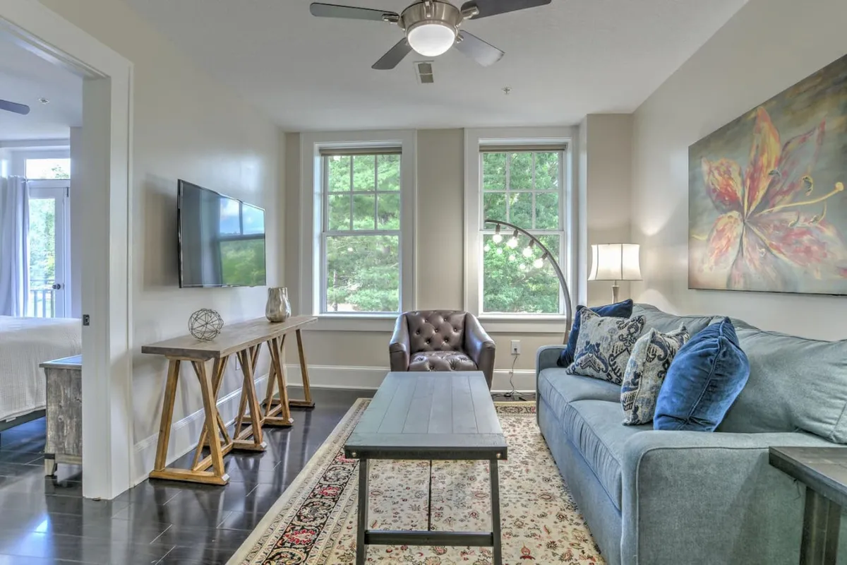 A welcoming living room featuring a sofa adorned with decorative pillows, artwork hung on the wall, a wooden coffee table, and a chestnut-colored armchair. Large windows allow natural light to flood the space, enhancing the cozy and inviting atmosphere.
