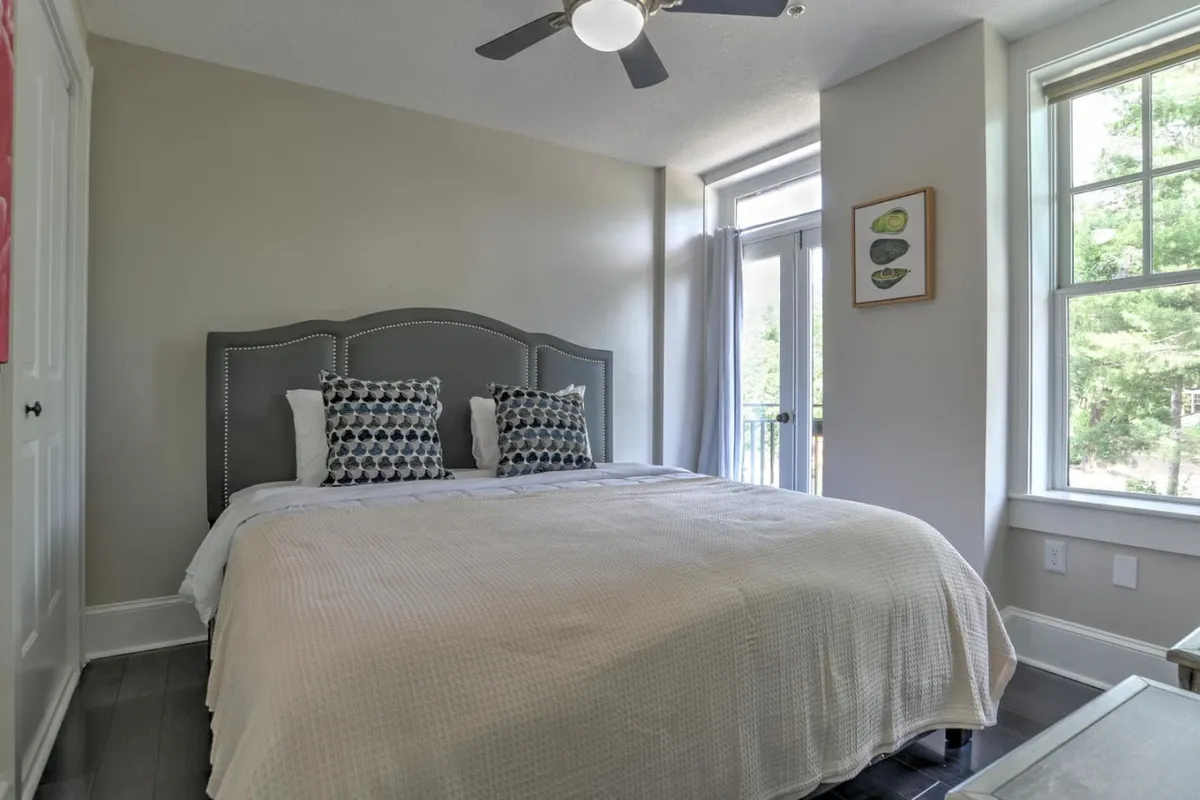 A bright, serene bedroom featuring a neatly made bed with decorative pillows, a large window flooding the space with natural light, and tasteful artwork adorning the wall. The room feels airy and restful, ideal for relaxation.