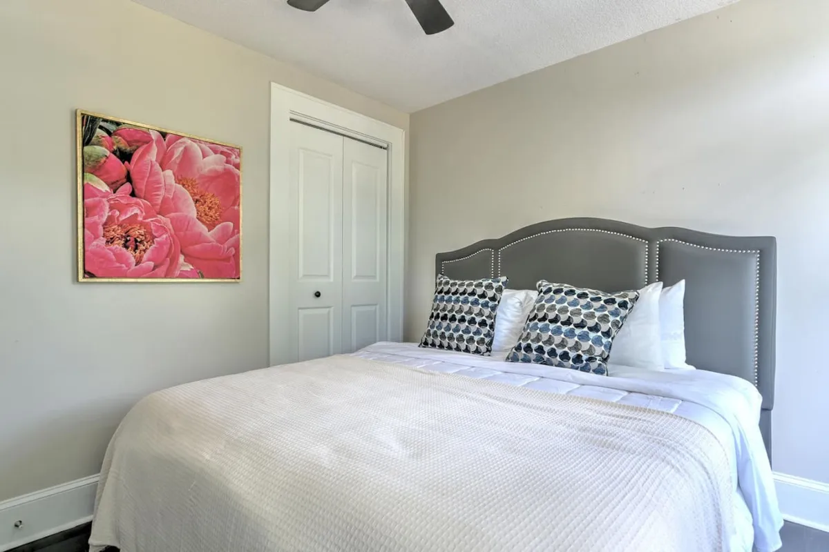 A cozy, sunlit bedroom is shown with a charming bed adorned with decorative pillows and framed floral artwork above. The soft lighting and inviting textures create a warm, intimate setting ideal for relaxation. Elements like the closet door and art add to the historic, character-filled ambiance described in the section.